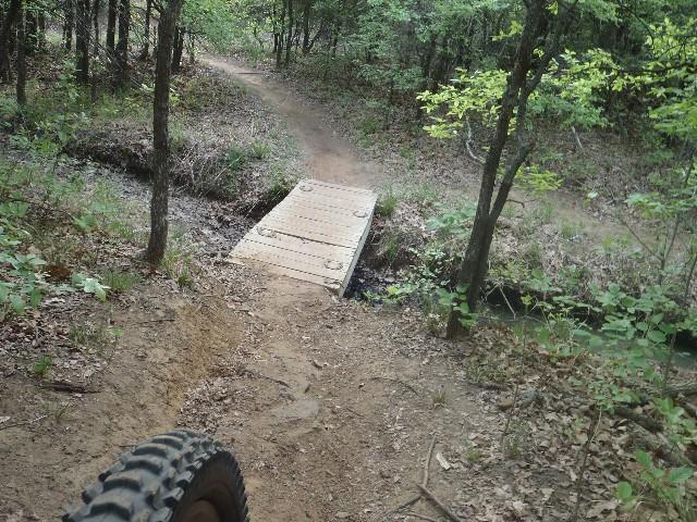 A narrow dirt path winding through a forest, featuring a wooden bridge crossing over a small creek. The foreground shows part of a tire, indicating the view from a bicycle. Lush greenery and trees line the trail, creating a serene natural setting. Horseshoe mountain bike trail.