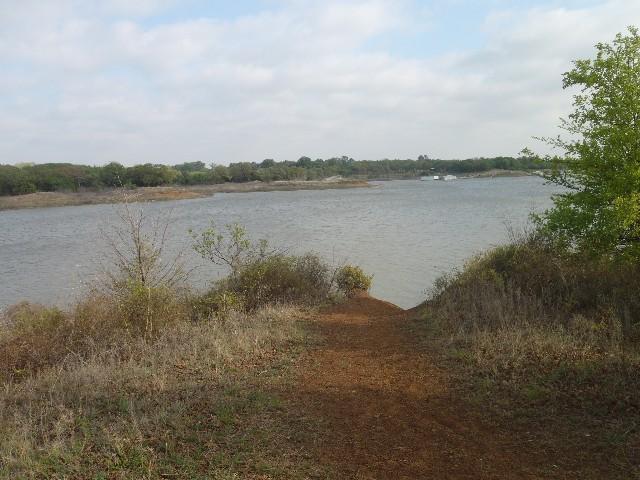 A scenic view of a calm lake surrounded by greenery, with a dirt pathway leading towards the water. The sky is partly cloudy, and the landscape features patches of grass, small shrubs, and trees in the background. Horseshoe mountain bike trail.
