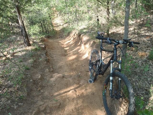 A mountain bike parked beside a sandy dirt trail surrounded by trees, indicating a forested area suitable for biking. The trail winds into the distance, showcasing natural terrain and greenery. Northshore Trail mountain bike trail.
