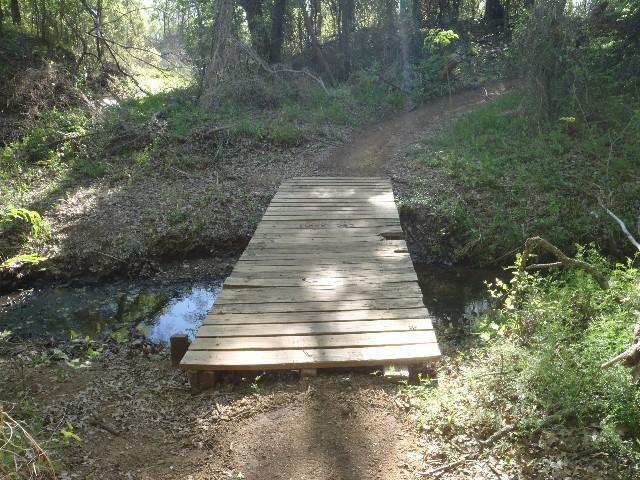 Wooden footbridge crossing a small creek, surrounded by lush greenery and sunlight filtering through trees. A dirt path leads up to the bridge on one side, while a continuation of the trail is visible on the other side. Northshore Trail mountain bike trail.