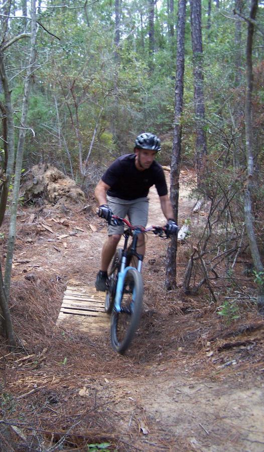 A mountain biker rides along a narrow dirt trail surrounded by trees, navigating a wooden bridge on the path. The rider, wearing a helmet and gloves, appears focused as they maneuver over the bridge in a forested area. Games Loop mountain bike trail.