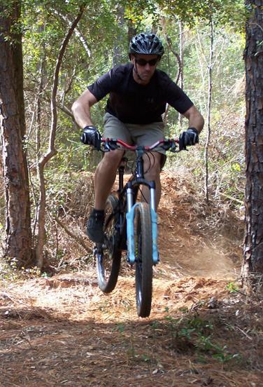 A cyclist in a black helmet and gloves is mid-air while performing a jump on a mountain bike along a dirt trail surrounded by trees. Dust is being kicked up from the ground as he descends a slope. The scene captures the excitement and skill involved in mountain biking. Games Loop mountain bike trail.