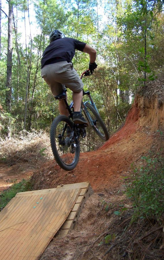 A mountain biker executes a jump off a wooden ramp on a dirt trail, surrounded by trees and lush vegetation. The rider is in mid-air, showcasing the excitement of off-road biking. Games Loop mountain bike trail.