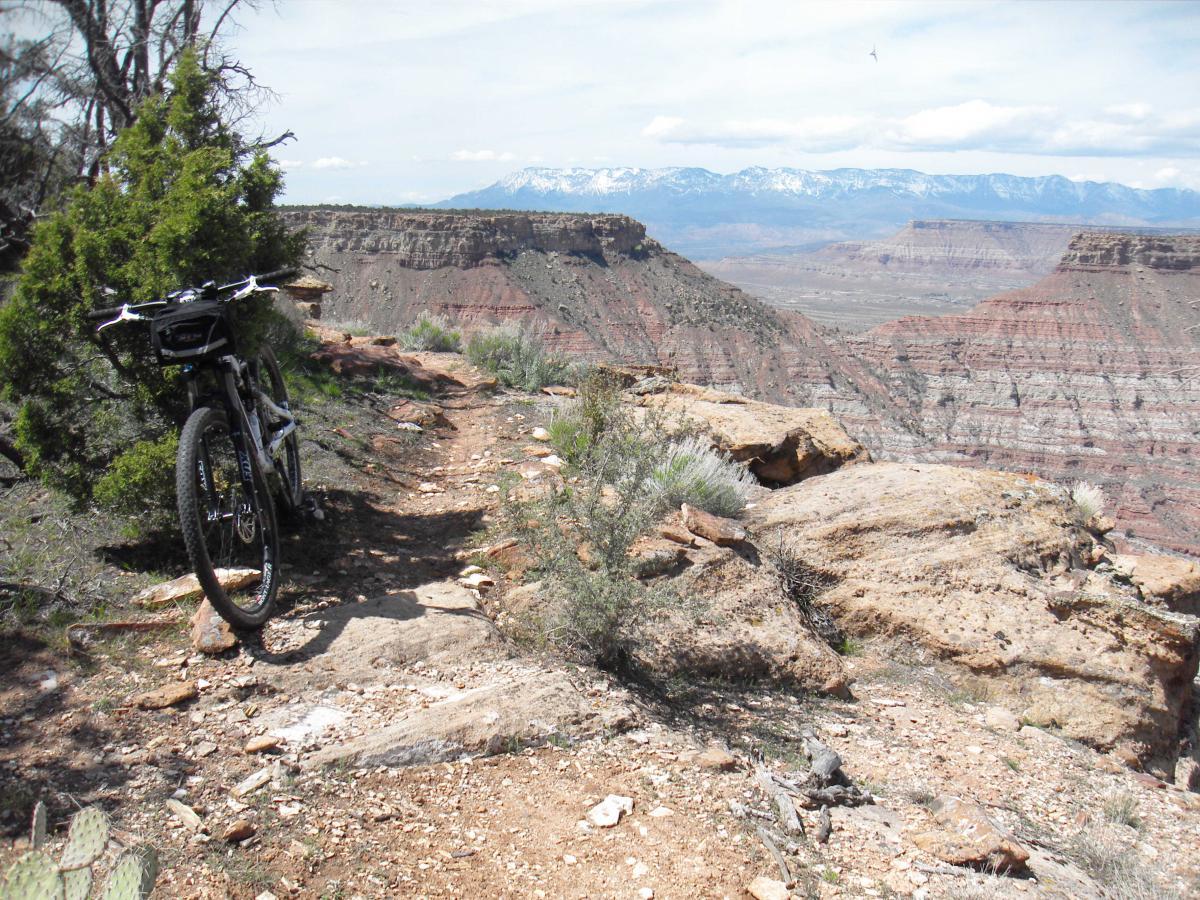 A mountain bike parked on a rugged dirt trail overlooking a vast canyon with layered rock formations and distant snow-capped mountains under a partly cloudy sky. Gooseberry Mesa mountain bike trail.