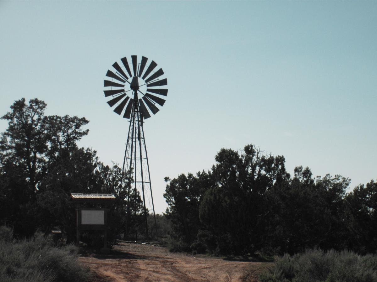 A windmill stands tall against a clear sky, surrounded by dense greenery. In the foreground, a wooden information board is partially visible, set along a dirt path. The scene evokes a sense of rural tranquility and connection to nature. Gooseberry Mesa mountain bike trail.