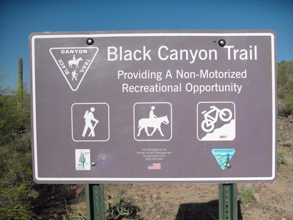 A sign for the Black Canyon Trail, indicating a non-motorized recreational area. The sign features symbols for hiking, horseback riding, and biking. The background includes desert vegetation and a clear blue sky. Black Canyon Trail - Little Pan Loop mountain bike trail.