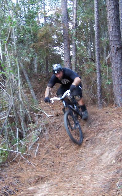 A mountain biker navigating a narrow, winding trail through a wooded area, leaning forward and ascending a steep incline. The rider is wearing a black helmet and gear, with trees and foliage in the background creating a natural setting. Games Loop mountain bike trail.