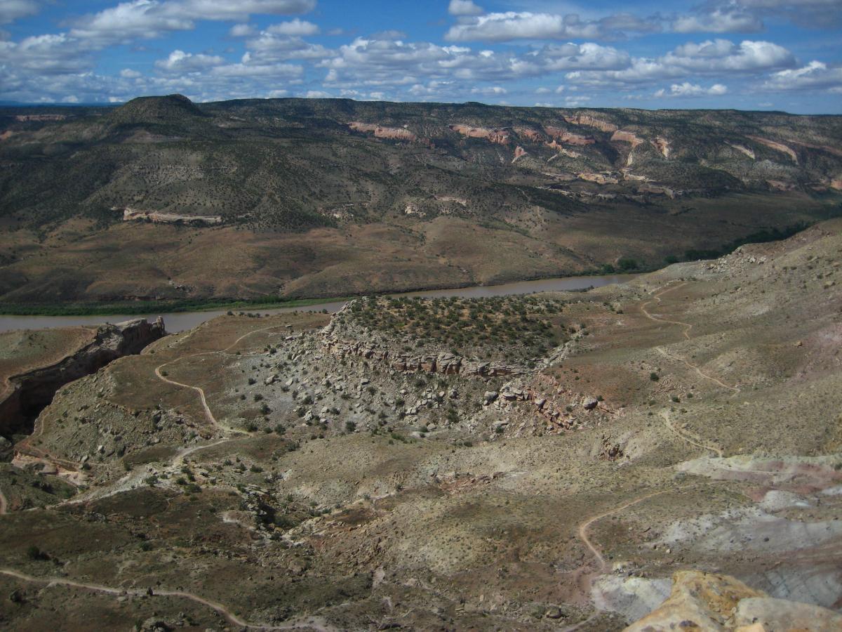A panoramic view of a rugged landscape featuring rolling hills, a winding river, and scattered vegetation under a partly cloudy sky. The terrain shows a mix of rocky outcrops and dirt paths, highlighting the natural beauty and varying topography of the area. Mary's Loop / Horsethief Bench mountain bike trail.