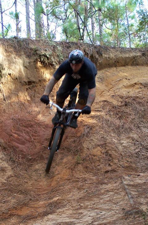A cyclist riding down a dirt slope in a wooded area, with trees in the background. The rider is wearing a helmet and has a focused expression, navigating the uneven terrain on a mountain bike. Games Loop mountain bike trail.
