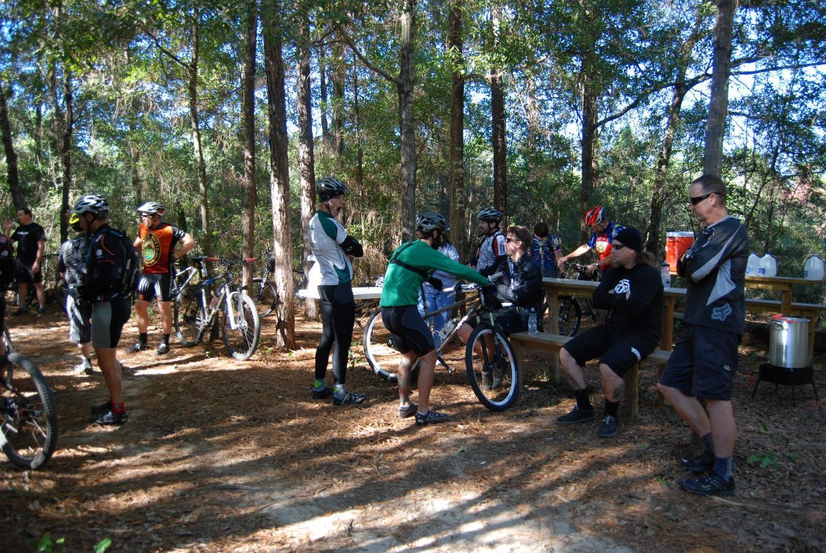 A group of mountain bikers gathered in a wooded area, taking a break. Some are chatting and socializing, while others are adjusting their bikes. The scene features various biking gear and helmets, with several bicycles parked nearby. Sunlight filters through the trees, casting dappled shadows on the ground. A wooden bench can be seen in the background, along with containers for refreshments. UWF Mountain Bike Trails mountain bike trail.