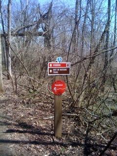 A wooden trail sign in a wooded area, displaying information about a nature trail. The sign features directional arrows, a blue symbol indicating the trail, and the name "Vardon" prominently displayed. Surrounding the sign are bare trees and underbrush, typical of a natural outdoor setting. MoMBA @ Huffman MetroPark mountain bike trail.