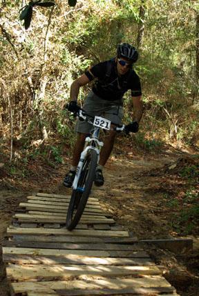 Mountain biker navigating a wooden plank bridge on a forest trail, wearing a helmet and jersey, with a race number displayed on his bike. UWF Mountain Bike Trails mountain bike trail.