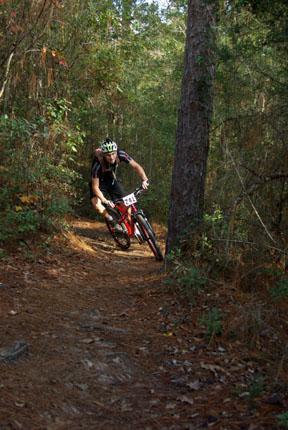 A mountain biker maneuvering sharply on a dirt trail through a wooded area, surrounded by trees and foliage. The rider wears a helmet and cycling gear, and the bike is red with a number plate. UWF Mountain Bike Trails mountain bike trail.