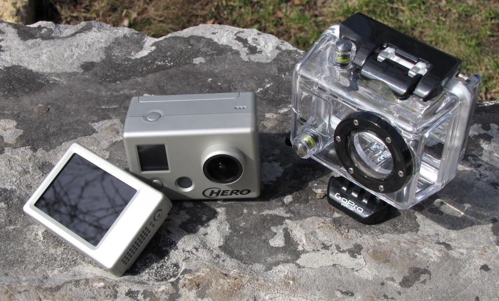 A silver action camera labeled "HERO" sitting on a stone surface, accompanied by a small display screen and a clear waterproof housing case. The background features grass and a blurred natural setting.