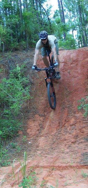 A mountain biker descending a dirt slope surrounded by trees and greenery, wearing a helmet and protective gear. Games Loop mountain bike trail.