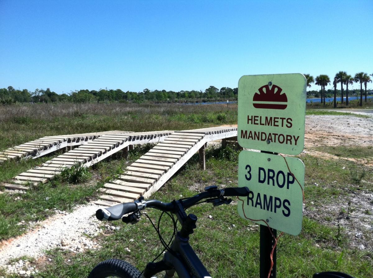A mountain bike positioned beside a sign stating "Helmets Mandatory" and "3 Drop Ramps," with wooden ramps visible in the background, set against a clear blue sky and grassy landscape. Quiet Waters Park mountain bike trail.