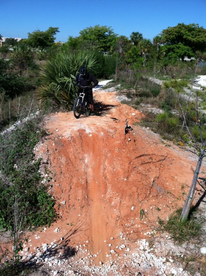 A person wearing a helmet and black clothing stands next to a mountain bike on a dirt jump. The jump is made of reddish-brown dirt with a steep incline, surrounded by sparse vegetation and tall grass under a clear blue sky. Quiet Waters Park mountain bike trail.
