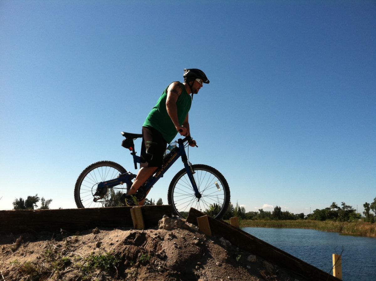 A person wearing a green tank top and a helmet balances on a blue mountain bike at the edge of a wooden ramp overlooking a body of water. The scene is set under a clear blue sky with a few trees visible in the background. Quiet Waters Park mountain bike trail.