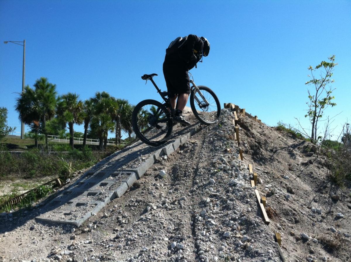 A mountain biker balancing on a steep dirt ramp, surrounded by palm trees and blue skies, preparing to ride down the slope. Quiet Waters Park mountain bike trail.
