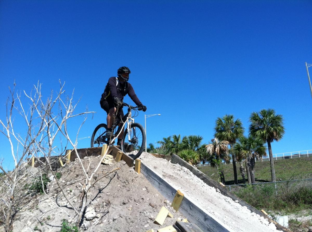 A mountain biker navigating a dirt ramp, positioned on top of a slope with wooden supports. The sky is clear and blue, and palm trees are visible in the background, indicating a sunny outdoor location. Quiet Waters Park mountain bike trail.