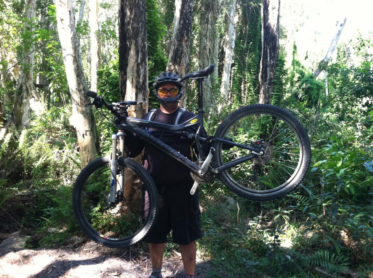 A person wearing a helmet and sunglasses holds a mountain bike above their head in a wooded area. The background features tall trees and lush green foliage, suggesting a natural outdoor setting suitable for biking. The person is dressed in casual biking attire, ready for an adventurous ride. Okeeheelee Park / Pinehurst / Green Acres Freedom Park mountain bike trail.