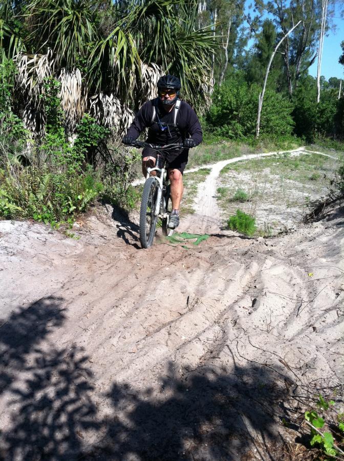 A mountain biker riding on a sandy trail surrounded by lush greenery and palm trees. The cyclist is wearing a helmet and sunglasses, navigating a slight incline on the path. Okeeheelee Park / Pinehurst / Green Acres Freedom Park mountain bike trail.