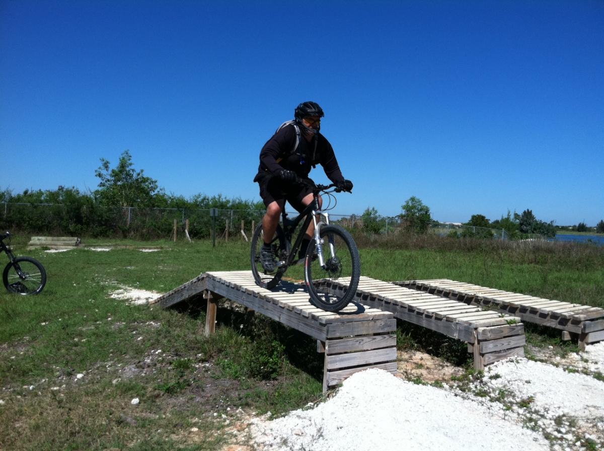 A mountain biker riding across a wooden ramp in a grassy outdoor area on a sunny day. In the background, there is lush greenery and a clear blue sky. Another bike can be seen to the left, partially obscured. Quiet Waters Park mountain bike trail.