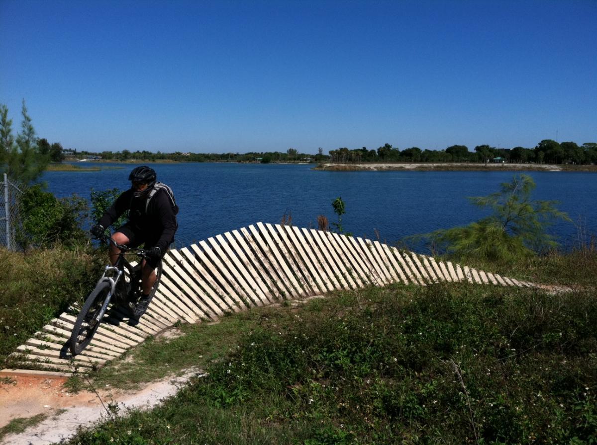 A mountain biker navigates a wooden sloped trail alongside a calm lake on a clear, sunny day. Lush greenery surrounds the biking path, and a blue sky complements the serene water in the background. Quiet Waters Park mountain bike trail.