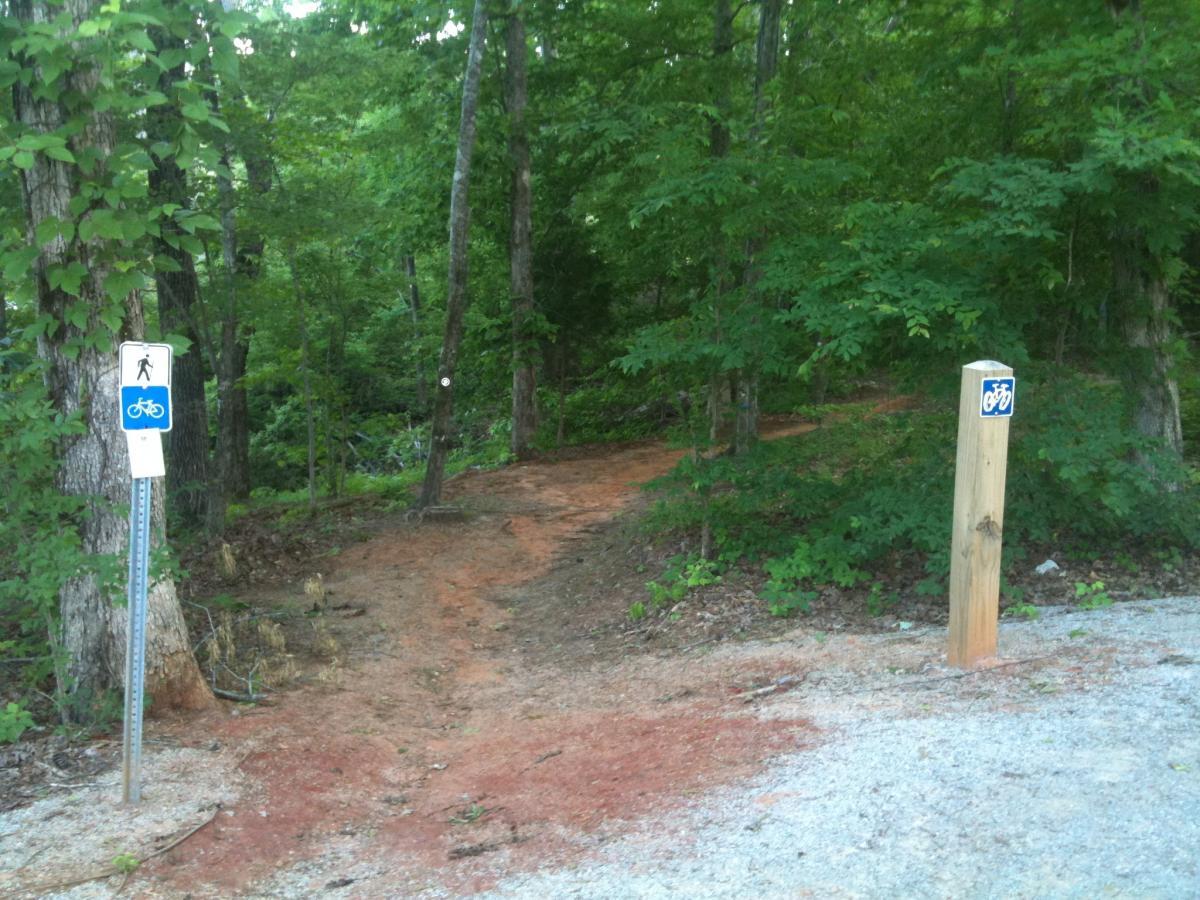 A pathway in a wooded area with signs indicating trail use for pedestrians and bicycles. The path is partially visible, surrounded by greenery and trees. One sign shows a hiker icon, while the other indicates a bicycle. Bonita Lakes mountain bike trail.