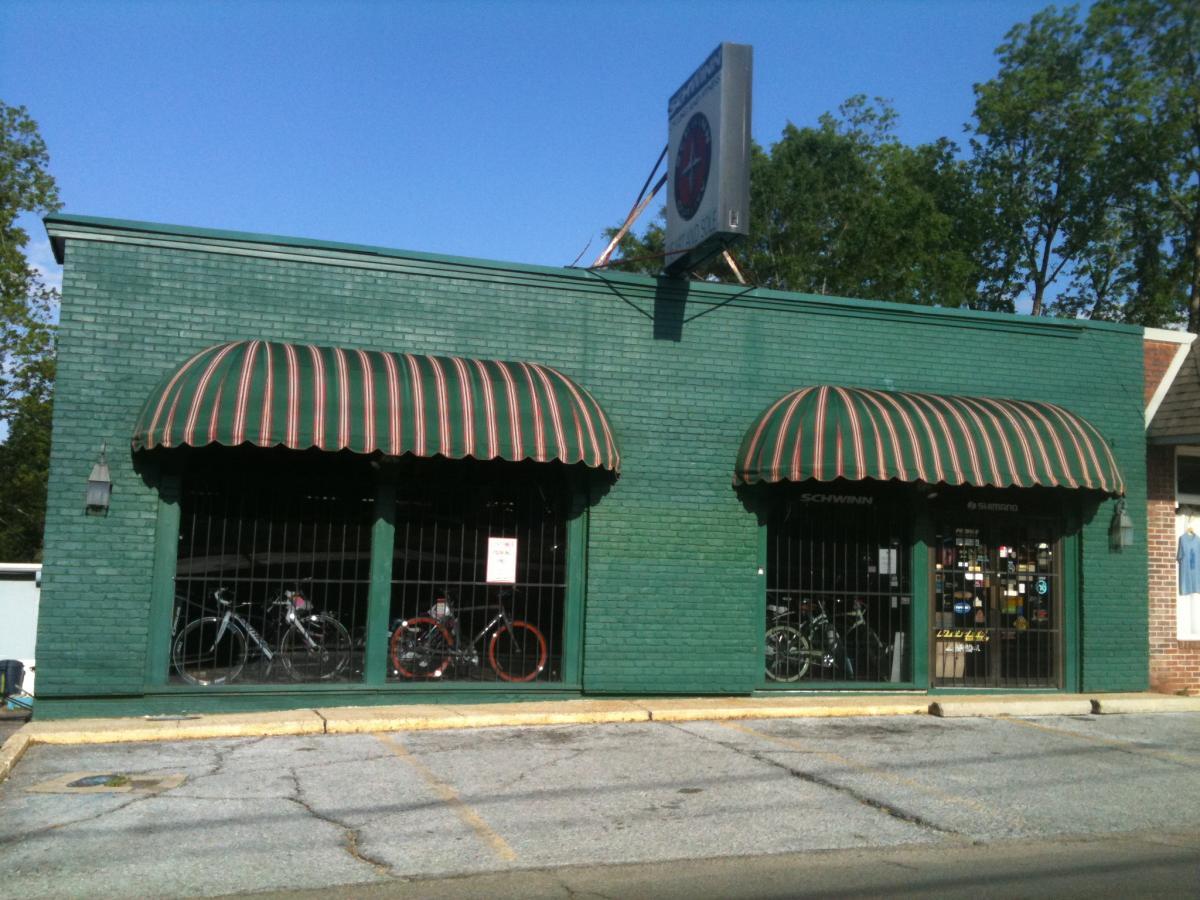 A green brick storefront with two striped awnings, showcasing bicycles behind gated windows. A sign on the roof displays a logo. The parking lot in front features cracked pavement, and trees are visible in the background under a clear blue sky.