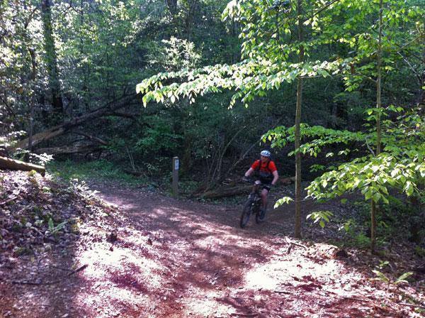 A person riding a mountain bike on a dirt trail surrounded by lush green trees and vegetation, with sunlight filtering through the leaves. The trail appears to be winding, with a signpost visible in the background. Overmountain Victory Trail mountain bike trail.