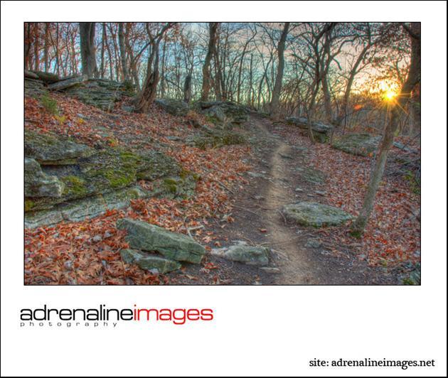 A winding dirt path meanders through a wooded area, bordered by rocks and scattered autumn leaves. The sun is setting in the background, casting a warm glow through the trees. Swope Park Trail mountain bike trail.