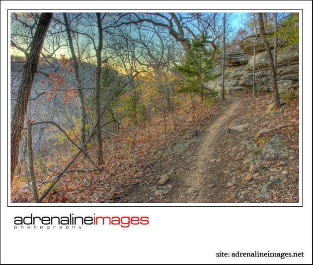 A winding dirt trail surrounded by bare trees and rocky formations, set in a peaceful, natural landscape during the late afternoon. The scene captures the tranquility of the outdoors with soft, warm lighting filtering through the branches. Swope Park Trail mountain bike trail.