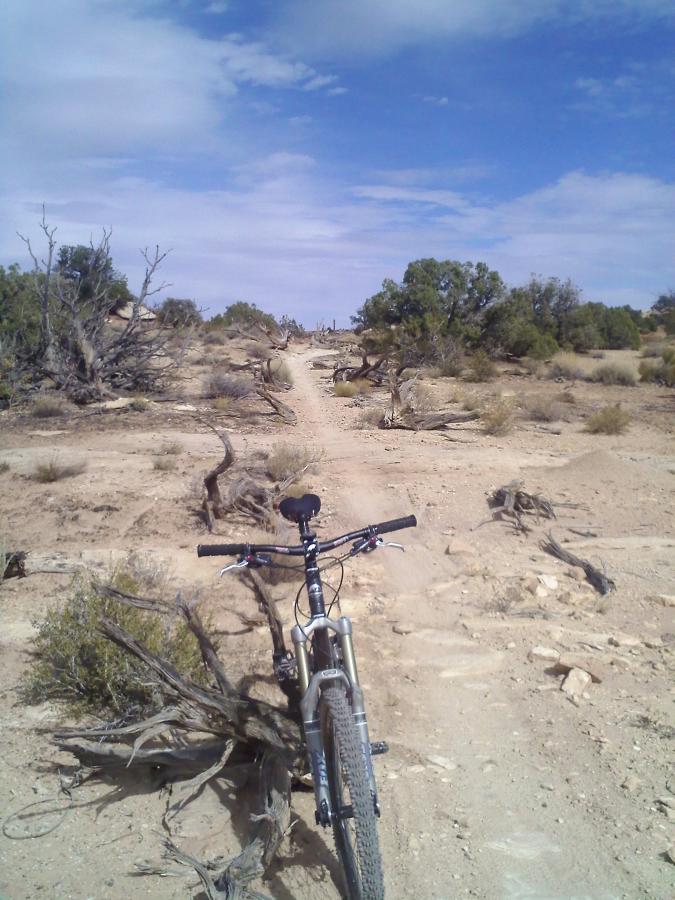A view of a dirt mountain biking trail stretching into the distance, with a bike positioned in the foreground. The landscape is arid, featuring sparse vegetation and rugged terrain, under a partly cloudy blue sky. Klondike Bluffs mountain bike trail.