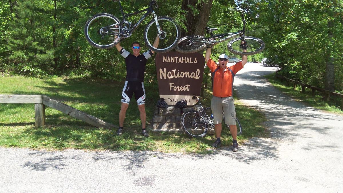 Two cyclists celebrating at the Nantahala National Forest sign. They are holding their bikes overhead, smiling and posing for the camera. The scene is surrounded by greenery, with a path leading into the forest visible in the background. Tsali Recreation Area mountain bike trail.