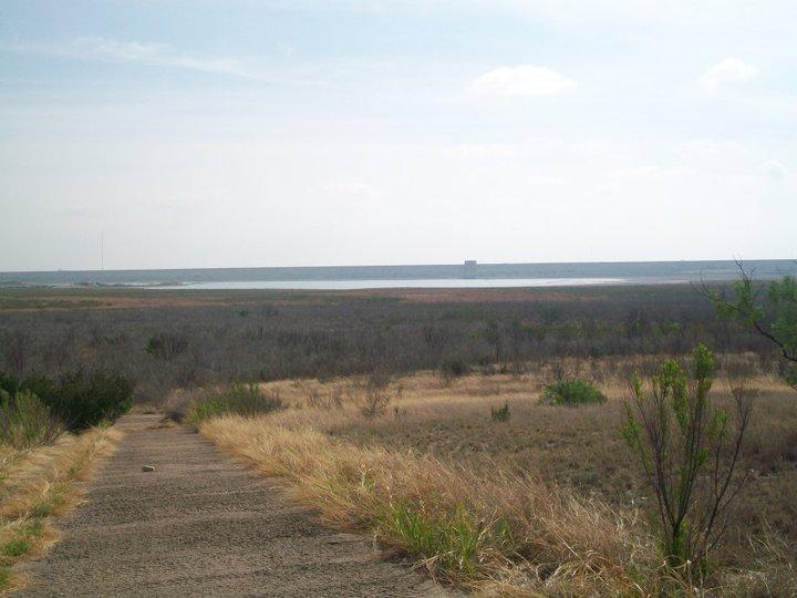 A pathway leading down to a large body of water, surrounded by dry grass and sparse vegetation under a cloudy sky. The horizon features a calm lake with distant structures visible on the water's edge. San Angelo Trail mountain bike trail.