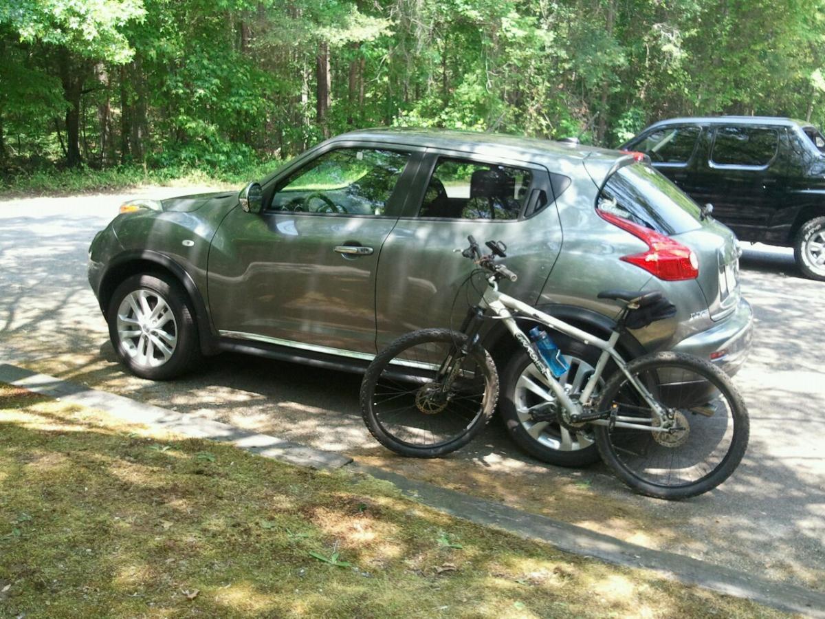 A silver SUV parked alongside a quiet road, with a bicycle leaning against its side. The surrounding area features lush green trees and grass, indicating a natural setting. Another vehicle is visible in the background. Paynes Creek mountain bike trail.