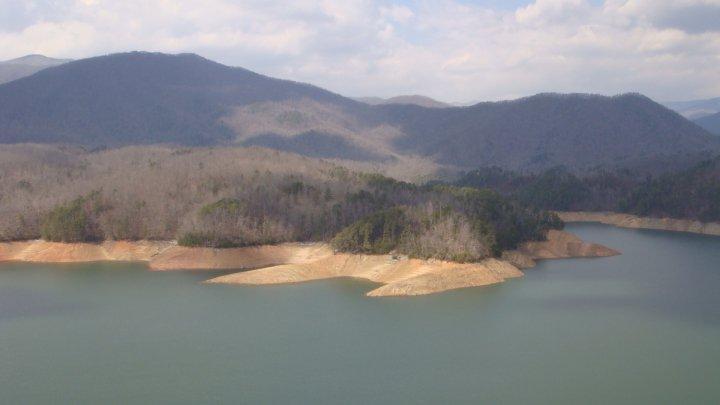 A scenic view of a calm lake surrounded by rolling hills and mountains, with patches of bare trees along the shoreline and a cloudy sky overhead. The image captures the serene landscape with a mix of water and land, showcasing the natural beauty of the area. Tsali Thompson Loop mountain bike trail.