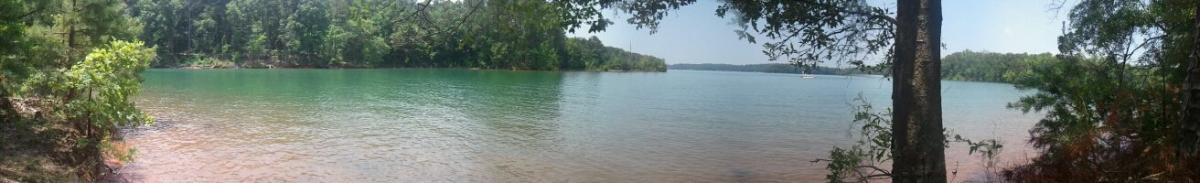 A panoramic view of a calm lake surrounded by lush green trees and vegetation on a sunny day. The water is a mix of turquoise and clear shades, with a hint of reddish-brown shoreline visible in the foreground. The scene conveys a peaceful natural environment ideal for relaxation or outdoor activities. Paynes Creek mountain bike trail.