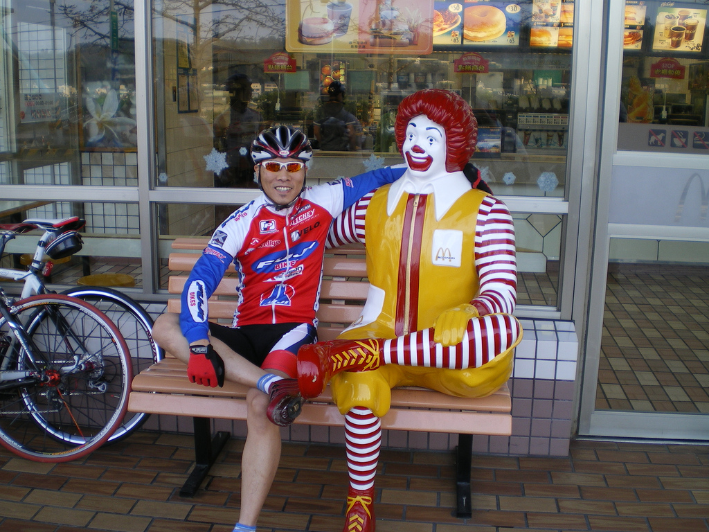 A cyclist in a colorful jersey sits on a bench beside a statue of Ronald McDonald outside a fast-food restaurant. The cyclist, wearing a helmet and sunglasses, smiles as he poses with the cheerful clown, who is dressed in his signature red and yellow outfit. A bicycle is parked nearby.