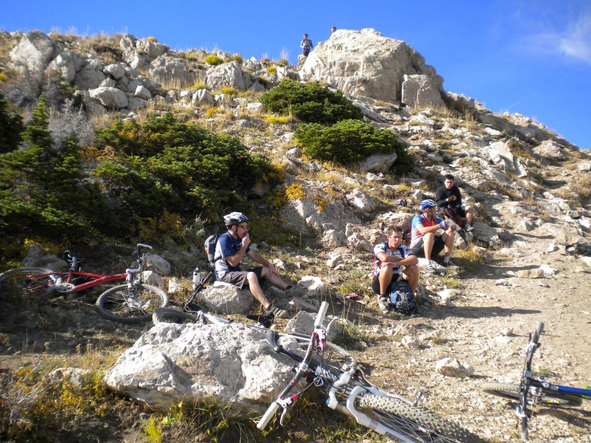 A group of mountain bikers resting on a rocky slope under a blue sky. Some bikes are scattered around them, and they appear to be enjoying a break amidst the natural landscape, characterized by grass and shrubs. Northern Skyline mountain bike trail.