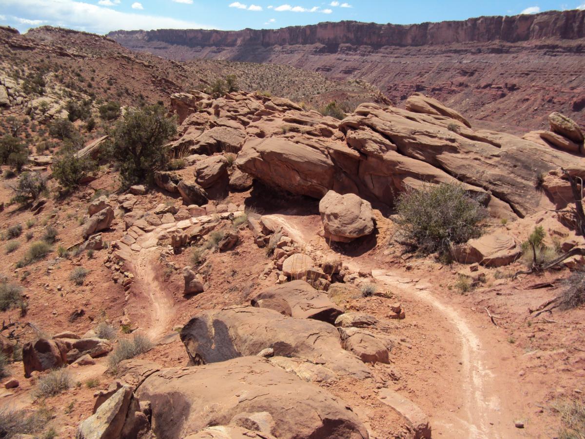 Winding dirt trail through rocky terrain in a desert landscape, with scattered shrubs and cliffs in the background under a blue sky with clouds. Deadman's Ridge mountain bike trail.