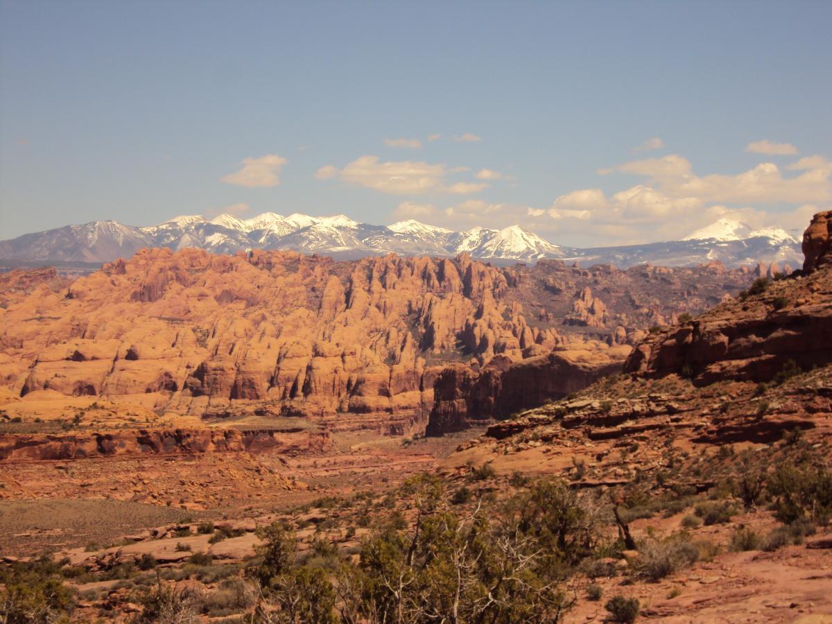 A panoramic view of a rugged landscape featuring towering reddish rock formations in the foreground and snow-capped mountains in the background under a clear blue sky. The scene includes patches of green vegetation scattered throughout the rocky terrain. Rockstacker mountain bike trail.