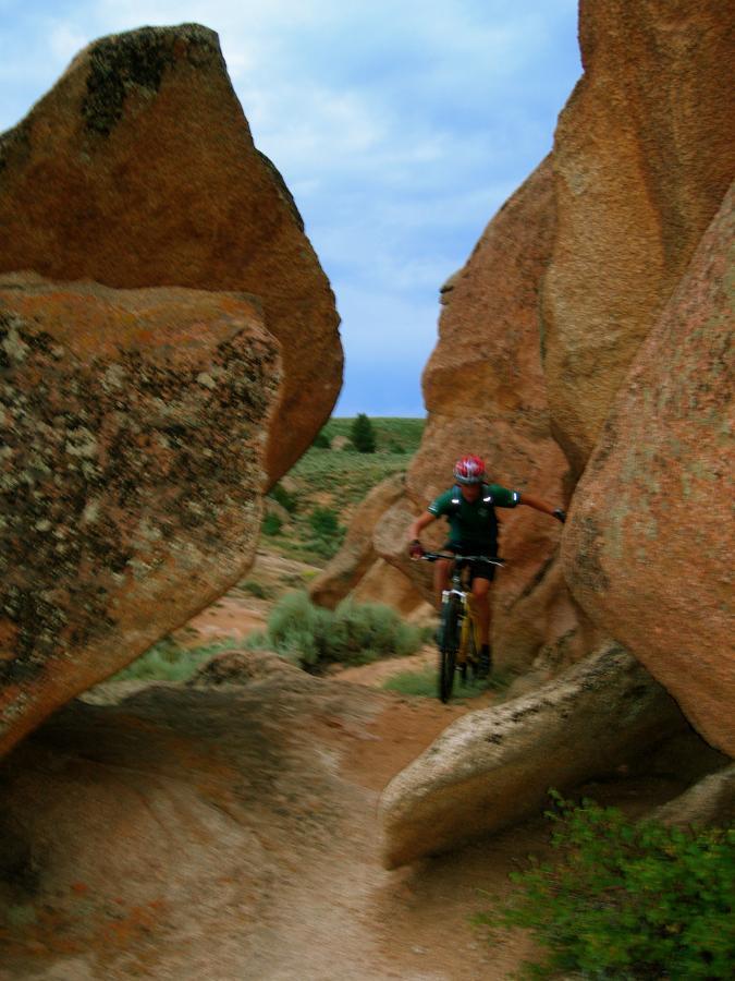A mountain biker navigating through a narrow passage between large boulders, set against a backdrop of rugged terrain and a cloudy sky. Hartman Rocks mountain bike trail.