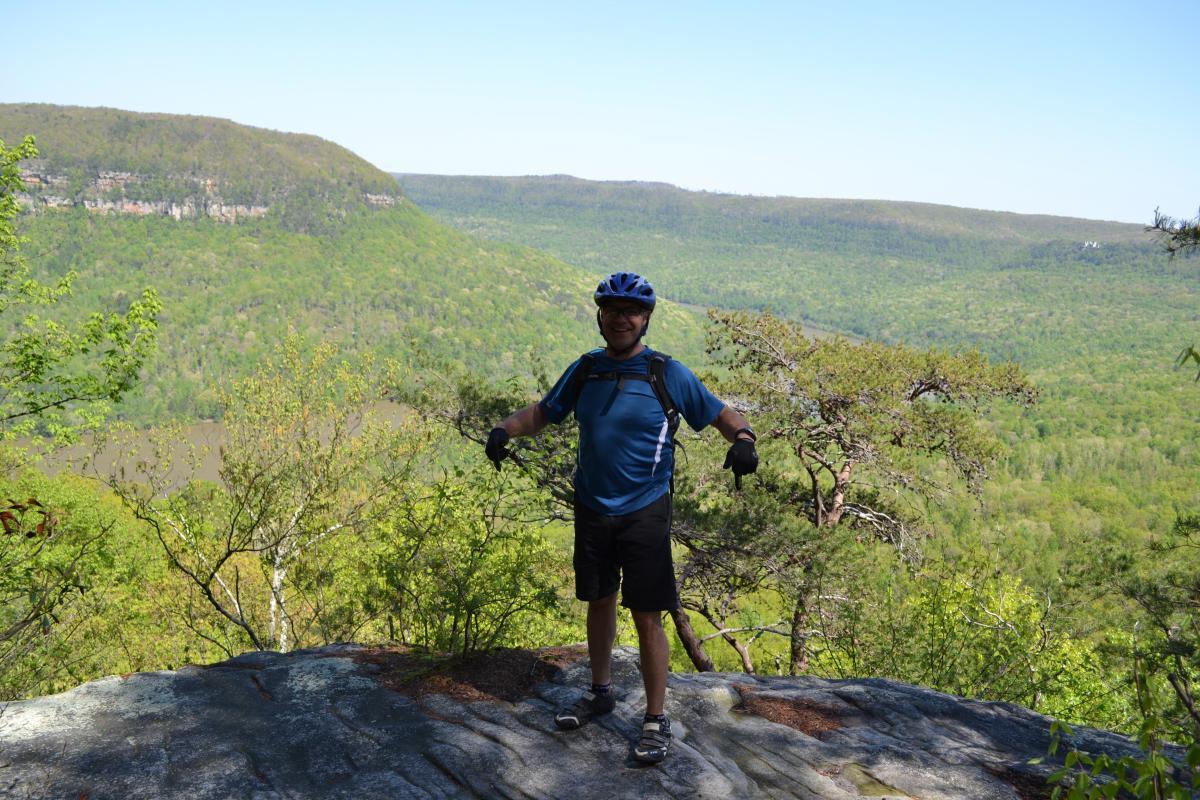 A person wearing a blue shirt, black shorts, and a helmet stands on a rocky outcrop, with their arms outstretched in excitement. They are surrounded by a lush green landscape, featuring rolling hills and trees under a clear blue sky. The scene captures the beauty of nature and conveys a sense of adventure. Raccoon Mountain Trail Network mountain bike trail.