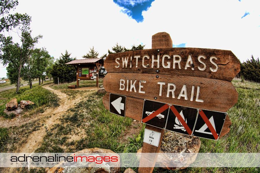 A wooden signpost labeled "Switchgrass Bike Trail" with directional indicators and icons for bike access, camping, and other trail regulations, set against a backdrop of green grass and trees under a cloudy sky. Switchgrass mountain bike trail.