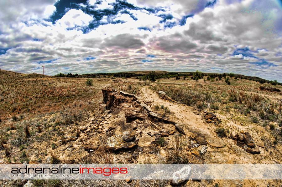 A panoramic view of a dry, rugged landscape under a partly cloudy sky. The foreground features exposed rocky terrain and patches of grass, while distant hills and trees create a natural backdrop. A power line can be seen on the left side of the image. Switchgrass mountain bike trail.
