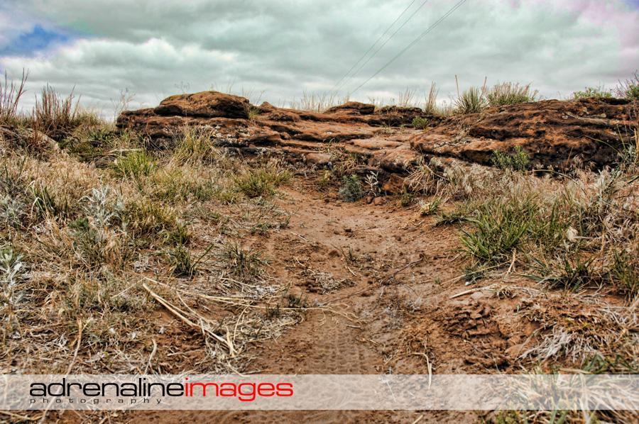 A dirt path surrounded by grass and rocky outcrops under a cloudy sky. The terrain appears rugged and natural, suggesting an outdoor setting, possibly in a rural or wild environment. Switchgrass mountain bike trail.