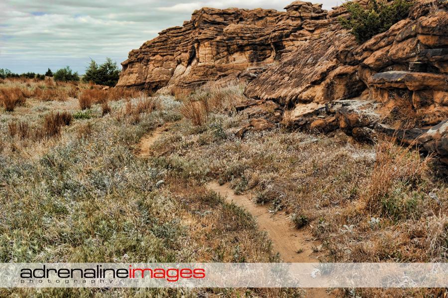 A winding dirt path leads through a rocky landscape with prominent cliffs on one side, surrounded by dry grass and sparse vegetation under a cloudy sky. Switchgrass mountain bike trail.