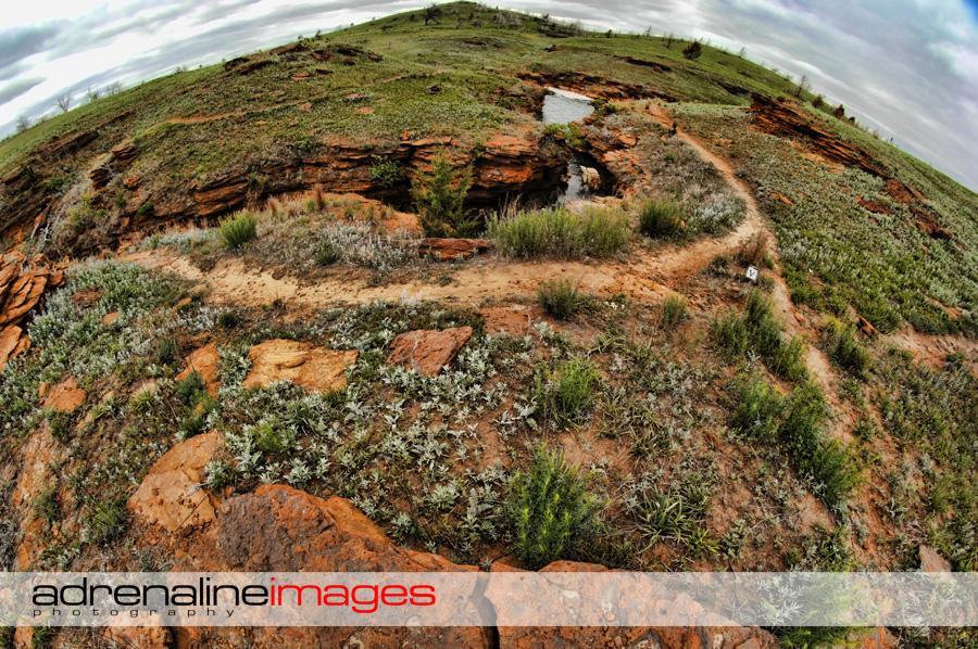A panoramic view of a rocky hillside with green vegetation and patches of grass. A winding dirt path is visible, leading through the landscape, which includes reddish rock formations and a small body of water. The sky is overcast with various shades of gray, suggesting a cloudy day. Switchgrass mountain bike trail.